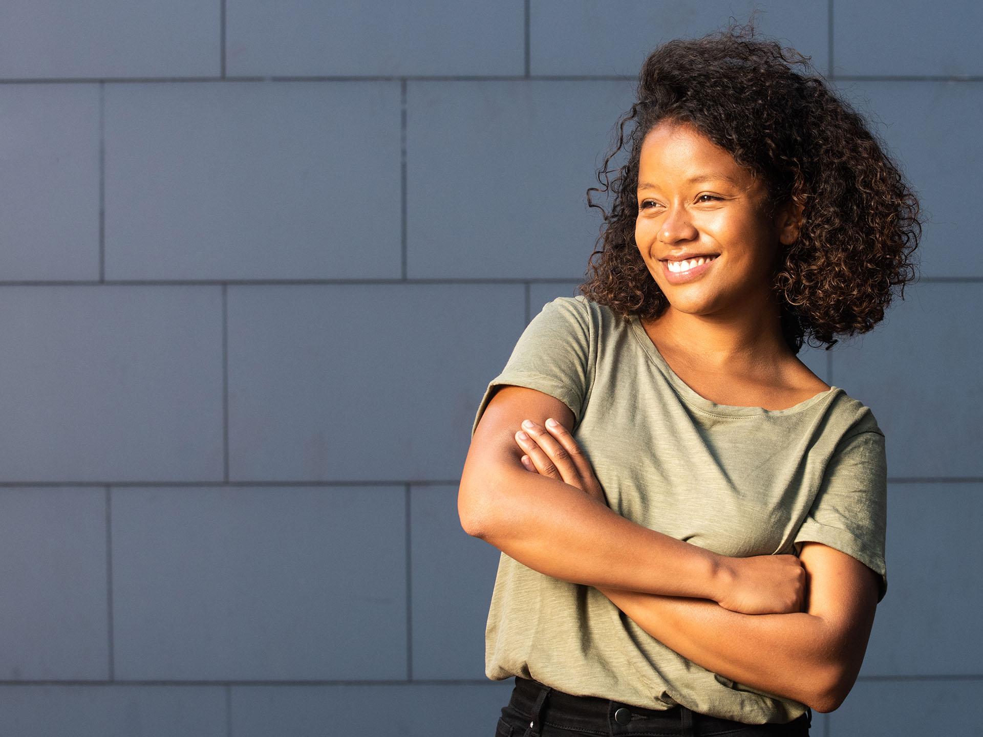 Happy young African American woman with arms crossed by gray background